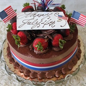 JC-006 Tiramisu topped with strawberries, chocolate shavings, mini American flags, and a "Happy July 4th" card. Red, white, and blue ribbon surrounds the cake for a festive patriotic look.