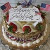 The JC-005 Carrot is a festive cake decorated with strawberries, whipped cream, chopped nuts, American flags, and a “Happy July 4th” sign. It sits on a gold tray with white ruffled fabric underneath.