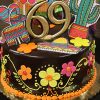 The CB-028B chocolate cake is decorated with colorful flowers, a sombrero, cactus, piñata, and a gold 69 topper for a festive Mexican-themed design.