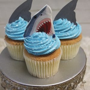 Three CC-027A cupcakes, gold with chocolate filling, each topped with blue frosting and decorative shark fins—one featuring a shark face—arranged on a silver ornate cake stand.