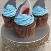 Three CC-027B chocolate cupcakes with white chocolate mousse filling, topped with blue frosting and shark-themed decorations, are displayed on a silver cake stand with hanging crystal accents.