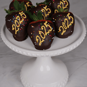 Six NY-013A Chocolate-covered strawberries decorated with yellow 2025 and colorful sprinkles are arranged on a white, beaded cake stand against a marble backdrop.