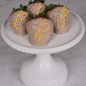 Four strawberries covered in NY-013B Milk chocolate with “2025” in yellow icing and colorful sprinkles, displayed on a white pedestal cake stand against a marble background.