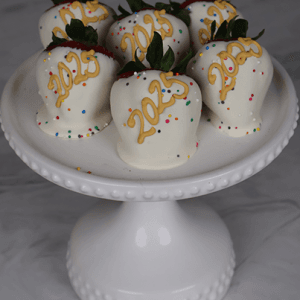Six strawberries dipped in NY-013C White Chocolate, topped with yellow "2023" decorations and colorful sprinkles, are displayed on a white cake stand against a gray marble background.