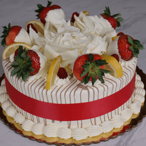 BD-003 Lemon/Raspberry is a round cake with white frosting, chocolate drizzle, strawberries, lemon slices, white chocolate curls, a red ribbon around the sides, piped cream at the base, and sits on a gold cake board.