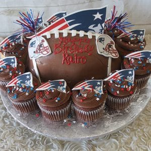 ST-002A Carrot Cake shaped like a football, decorated with New England Patriots logos, small American flags, star sprinkles, and "Happy Birthday Kairo" in red icing.