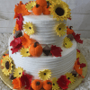 TC-008: A two-tiered white cake adorned with yellow sunflowers, red and orange autumn leaves, and small pumpkin-shaped accents, presented on a gold base.