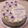 The BD-119 is a round cake with purple and white flowers, green edging, and "Congratulations Liz and Matt" in purple icing. It’s displayed on a silver board atop a checkered tablecloth.