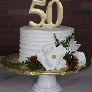The BD-132 is a white frosted cake on a pedestal stand with a gold "50" topper, white fondant flowers, pinecones, and green foliage, set against a brick wall background.