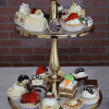 The AP-007 Assorted Mini Pastries Chef's Choice are elegantly arranged on a two-tier gold stand, featuring mini cakes topped with strawberries, chocolate, and cream, set against a brick wall and marble surface.