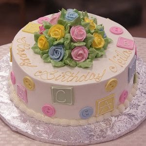 The BB-015 is a round birthday cake with pastel roses and yellow "Happy Birthday" text on top, plus pastel alphabet blocks and buttons on the sides. It sits on a silver foil-covered board.