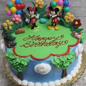 The SC-037 Mickey and Minnie cake features Mickey and Minnie toppers, colorful balloons, flower decor, green icing grass, "Happy Birthday" in red icing, blue sides with tree decorations.