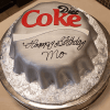 A round cake designed as a silver Diet Coke bottle cap, featuring "Happy Birthday Mo" in black icing. The BD-025 Diet Coke cake sits on a platter with a silver pattern.