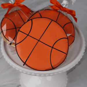 Three CE-002B Basketball-Shaped Cookies with orange icing and black lines are displayed on a white cake stand; two are wrapped in clear bags tied with orange bows.