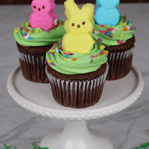 Three EA-048 Chocolate cupcakes with white filling, green frosting, colorful sprinkles, and pink, yellow, and blue marshmallow bunny candies sit on a white cake stand. Green paper grass decorates the surface below.