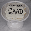 Two white frosted cookies with "GRAD" in black icing (Product: GC-0004) are displayed on a white decorative cake stand against a marble background.