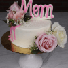 The MD-000C 6"cake petite One Size features white frosting, pink and white roses, baby’s breath, a pink “Mom” topper, gold cake board, white stand, and pink icing dripping down one side.
