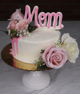The MD-000C 6"cake petite One Size features white frosting, pink and white roses, baby’s breath, a pink “Mom” topper, gold cake board, white stand, and pink icing dripping down one side.