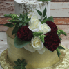 TC-011: A two-tiered cake with gold and white icing, adorned with white roses, dark red flowers, succulents, and greenery on an ornate gold board, set against a rustic wood background.