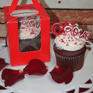 The VC-042 Individual Red Velvet cupcake, topped with white frosting, red heart sprinkles, and a red Love topper, sits on a white tray with rose petals, next to a matching cupcake in a red windowed box.
