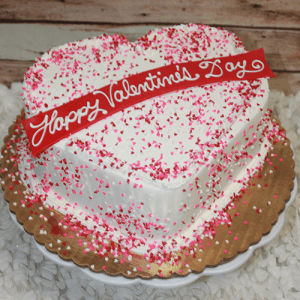 The VC-012 is a heart-shaped cake topped with white icing, pink and red sprinkles, and a red "Happy Valentines Day" banner in white script, presented on a gold cake board.