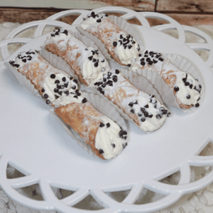 Six Cannolis with ricotta and chocolate chips are arranged on a white decorative plate, some in paper holders, all set on a white surface with a rustic wooden background.