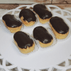 Six AP-045B Éclair pastries topped with glossy chocolate glaze are arranged on a white platter; two sit in white paper liners, while the others rest directly on the plate.