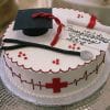 The GC-051 is a round cake topped with a graduation cap, diploma, stethoscope, red medical cross, piped heartbeat line, and "Congratulations Lucy 2016" written in icing.