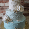 TC-015: Two-tiered cake with textured blue and white frosting, adorned with white sugar flowers, gold starfish, and white seashells on a gold cake board, set against a rustic wooden background.