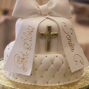 Product TC-021: White fondant cake with a large bow, gold cross, quilted design, "God Bless" and "Sonia" in gold on two ribbons, presented on a gold cake board.