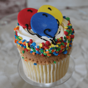 The CC-119 Gold cupcake features chocolate mousse filling, white frosting, colorful round sprinkles, and three balloon-shaped decorations in red, blue, and yellow. It’s presented on a glass plate.