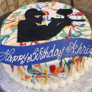 The CB-017A is a round birthday cake featuring colorful paint splatters, a black silhouette of a person with a paintball gun on top, and a blue "Happy Birthday Ashir" banner in white script.