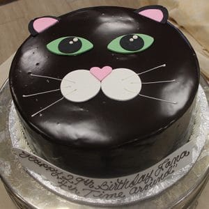 The HW-013 is a round chocolate cake decorated as a black cat’s face with green eyes, pink ears, a pink heart-shaped nose, white whiskers, and a birthday message on the base.