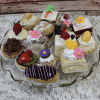 The EA-000A is a glass platter showcasing assorted mini desserts—cakes, tarts, cream puffs, and eclairs—decorated with fresh fruit, colorful icing, and edible flowers on a white textured surface.
