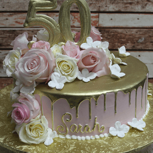 BD-057E Gold Pink champagne frosting cake, round with gold drip, pink and white roses, white flowers, large gold "50" topper, and "Sarah" in gold on the side. Presented on a gold board.