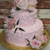 The TC-017 is a two-tier pink cake with white pearls, topped with pink roses and green leaves, presented on a gold cake board against a rustic wooden background.