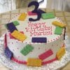 CB-004C is a round birthday cake featuring colorful toy brick-shaped pieces, pink piping, and a large purple number 3 on top, with "Happy Birthday Marisa" written in pink icing.