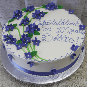 The BD-060E Periwinkle Hydrangeas is a round white cake adorned with purple flowers, green leaves, and a purple ribbon. "Congratulations on 100 years Dottie!" is written in purple icing.