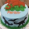 CB-014A is a round blue birthday cake decorated with green grass accents, black dinosaur silhouettes, "Parker" in orange icing, and topped with an orange dinosaur and a number 3.
