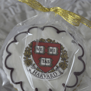 The GC-080K Harvard is a round cookie with scalloped white icing and maroon accents, featuring the Harvard University crest. It's wrapped in clear plastic and tied with a gold ribbon.