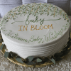 The bb-067 cake features a round, white frosted design with green and yellow floral accents and "baby IN BLOOM Welcome Baby Wesley" written in matching icing. It sits on a gold board with greenery at the base.