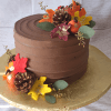 BR-053 is a chocolate frosted cake adorned with artificial autumn leaves, pinecones, and mini pumpkins on a gold cake board, showcased against a white textured background.