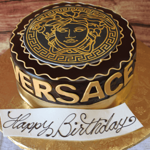 The BD-033 Designer Ganache/Gold is a round chocolate cake with the gold Versace logo and Medusa head on top, Versace lettering on the side, and a white "Happy Birthday" card in elegant script.