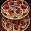 A two-tiered gold tray holds Fruit Tarts, each in a white paper cup and topped with strawberries, blueberries, raspberries, and kiwi slices. The tray rests on a dark wooden surface.