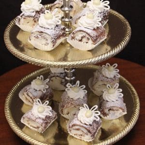 On a two-tiered gold tray, Marble Roulades—small rolled pastries with white icing, decorative swirls, and a white candy pearl—are dusted with powdered sugar. A dark background sets off the elegant sweets.