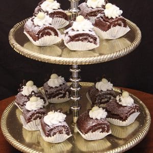 A two-tiered gold tray displays Oreo Roulades—chocolate Swiss roll slices with whipped cream and white chocolate, each in a white paper cup. The dark background sets off the desserts.