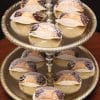 A two-tiered gold dessert stand displays Cannollis with Chocolate filling in white paper wrappers, each with chocolate-dipped ends and a dusting of powdered sugar.