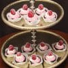 A two-tiered silver dessert stand displays twelve AP-032 Chocolate Raspberry Cups with pink frosting, each topped with a fresh raspberry and white chocolate drizzle, arranged neatly in white paper cups.