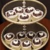 A two-tiered silver serving tray displays twelve Chocolate Cups in paper cups, each topped with white cream and drizzled chocolate, set against a dark background.