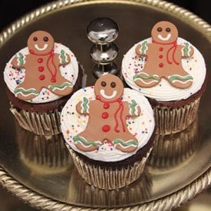 Three CH-066B chocolate cupcakes with white chocolate mousse filling, topped with white frosting, colorful sprinkles, and smiling gingerbread man cookies, are arranged on a round silver tray.
